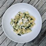 fusilli pasta with lemon zest, courgette and feta on a white plate, on a wooden table