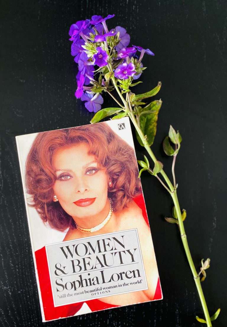 Sophia Loren's book Women and Beauty on a black table with a purple flower next to it